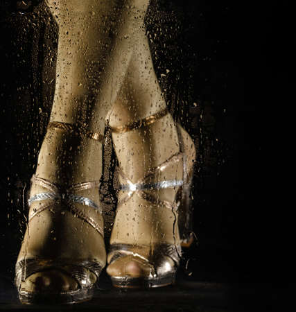 Female feet in high heeled sandals behind glass with water drops on black