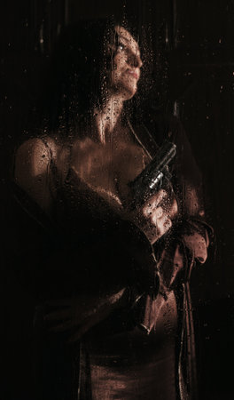 beautiful woman with a revolver in her hands behind a wet glass with raindropsの写真素材