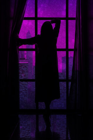 beautifu silhouette of a woman in a long peignoir at a large window on the background of the evening cityの写真素材
