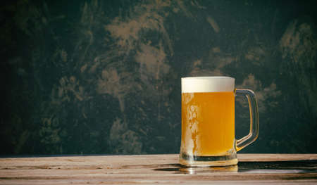 full mug with cold foamy beer on wooden table near wallの写真素材