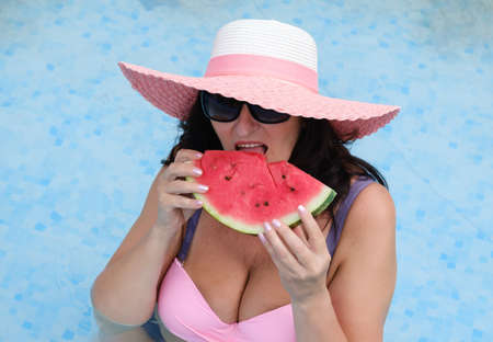 beautiful adult woman in a swimsuit, sunglasses and a summer hat holding a watermelon in the pool