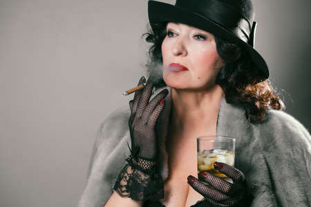 beautiful stylish woman in a fur coat, hat and gloves holds a glass with a drink and a cigarette in...