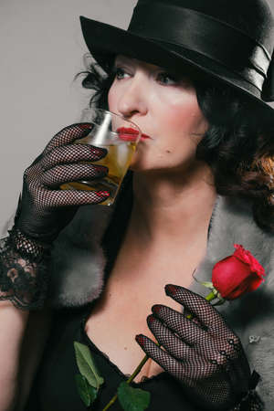 beautiful stylish woman in a fur coat, hat and gloves holds a glass with a drink and a red rose in...