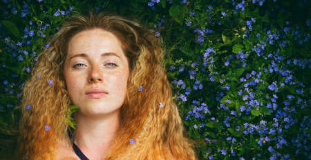 face of a beautiful red-haired woman with freckles on green grass among small beautiful purple...