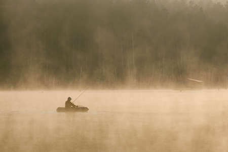 a fisherman in a boat catches fish in the early beautiful foggy morning and the forest in the...