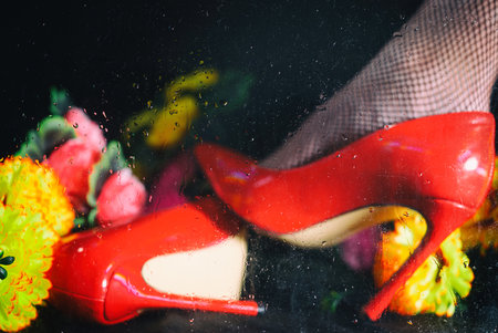 female leg in red stiletto heels and blooming spring flowers behind wet glass with water drops on black backgroundの写真素材
