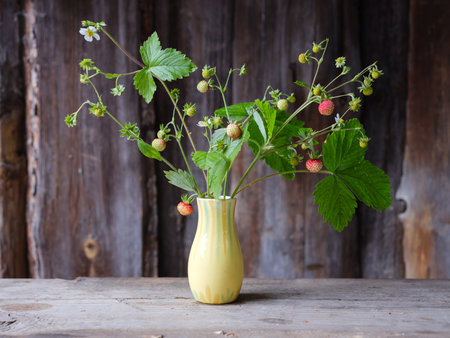 bouquet of strawberries with berries and flowers in a small yellow vase on a wooden table against a wooden wallの写真素材