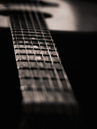 Black and white guitar with faint sepia, light hitting central part in focus, edges dark, neck extending into frame, background guitar softly blurred, moody and atmospheric composition emphasizing texture and light.の写真素材