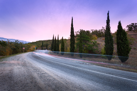 Mountain winding road passing through the forest with blurred car lights in motion with colorful sky at sunset in summerの写真素材