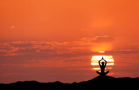 Sunset landscape with girl practicing yoga on the background of sun and red sky in the hill in summerの写真素材
