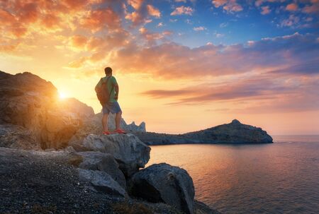 Beautiful summer landscape with standing man with backpack on the stone at the ocean against the colorful sky with clouds at sunset. Travel background. Sport, lifestyle. Tourismの写真素材