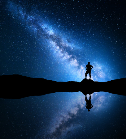 Milky Way. Night sky with stars and silhouette of a standing alone man on the mountain near the lake with reflection in water. Milky way and man on the hill. Galaxy and silhouette of a man. Universeの写真素材