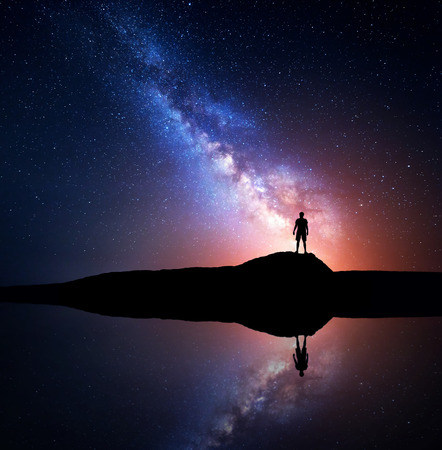 Milky Way. Night sky with stars and silhouette of a standing alone man on the mountain near the lake with reflection in water. Milky way and man on the hill. Galaxy and silhouette of a man. Universeの写真素材