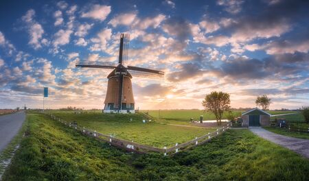 Windmill at sunrise in Netherlands. Beautiful old dutch windmill, green grass, fence  against colorful sky with clouds. Spring panoramic landscape in the morning in Holland. Rural scene. Travelの写真素材