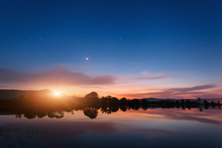 Mountain lake with moonrise at night. Night landscape with river, trees, hills, moon, stars and colorful blue sky with clouds reflected in water. Beautiful nature background in twilight. Travelの写真素材