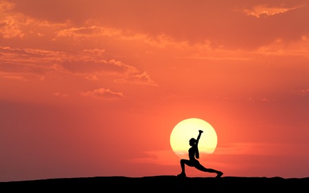 Silhouette of a standing sporty woman practicing yoga with raised up arms on the hill on the background of yellow sun and orange sky with clouds. Landscape with girl at sunset. Fitness and lifestyleの写真素材