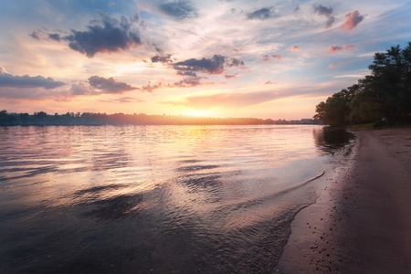 Colorful sunset at the river. Summer landscape with river, bright sky with clouds and sunlight reflected in water and trees on the beach. Nature background. Amazing view on the lake in the evening.の写真素材
