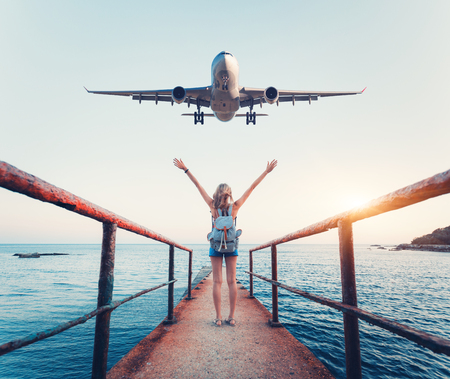 Airplane and woman at sunset. Summer landscape with girl standing on the sea pier with raised up arms and flying passenger airplane. Woman and landing commercial plane in the evening. Lifestyleの写真素材