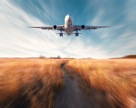 Airplane with motion blur effect. Landscape with flying passenger airplane and blurred blue sky with clouds, orange grass field with trail at sunset. Passenger airplane is landing. Commercial aircraftの写真素材