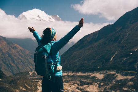 Standing young woman with backpack and raised up arms on the hill and looking on mountains. Landscape with happy girl, mountains, blue sky with clouds in autumn in Nepal. Travel. Trekking in Himalayasの写真素材