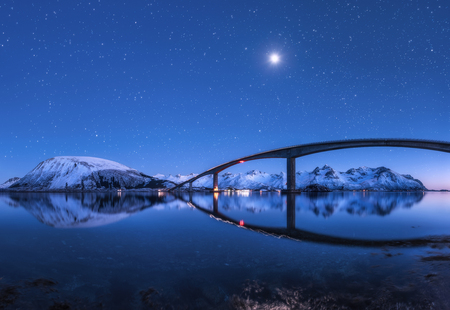 Amazing bridge and starry sky with beautiful reflection in water. Night landscape with bridge, snowy mountains, blue sky with moon and bright stars reflected in sea. Winter in Lofoten islands, Norway