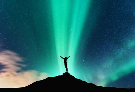 Aurora and silhouette of standing woman with raised up arms on the mountain in Norway. Aurora borealis and happy girl. Starry sky, green polar lights. Night landscape. Northern lights. Travelの写真素材