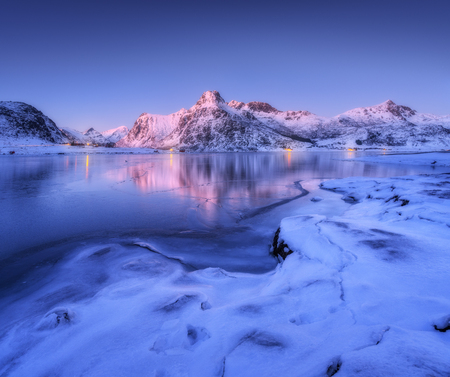 Frozen sea coast and beautiful snow covered mountains in winter at dusk. Beautiful fjord at night in Lofoten Islands, Norway. Nordic landscape with water, ice, rocks, buildings, illumination, blue skyの写真素材