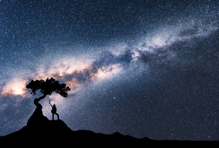 Milky Way and silhouette of woman under the tree growing from the rock on the mountain at night. Space background with starry sky, beautiful galaxy and girl. Scenery with Milky Way and people. Travelの写真素材