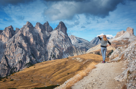 Young woman on the trail is looking on majestic mountains at sunset in autumn in Dolomites, Italy. Landscape with girl, cloudy sky, orange grass, high rocks and trees in fall. Travel in italian alpsの写真素材