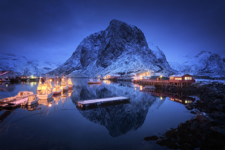 Fishing village with boats at night, Lofoten islands, Norway. Winter landscape with houses, pier, illumination, snowy mountains, sea, blue cloudy sky reflected in water at dusk. Norwegian rorbuerの写真素材