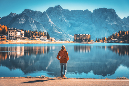 Young woman standing on the coast of Misurina lake at sunrise in autumn. Dolomites, Italy. Landscape with girl in red jacket, reflection in water, buildings, blue sky. Italian alps. Travel backgroundの写真素材