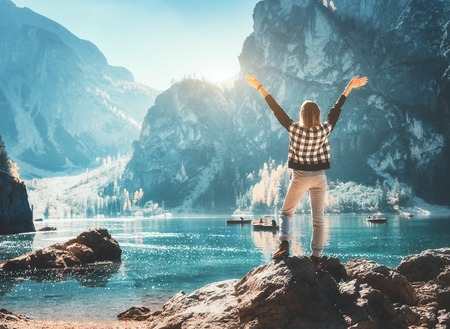 Happy young woman on the stone with raised up arms on the coast of Braies lake at sunset. Autumn in Dolomites, Italy. Landscape with girl, mountains, water with reflection, fall trees and blue skyの写真素材