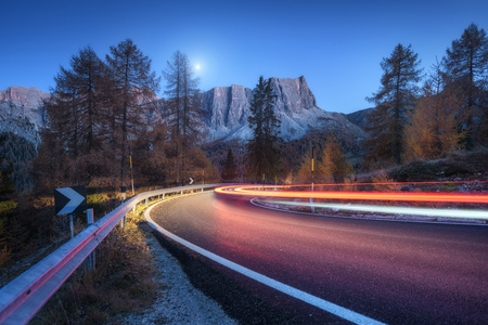 Blurred car headlights on winding road at night in autumn. landscape with asphalt road, light trails, mountains, trees, and blue sky with moon at dusk. Roadway in Italy. View with highway and rocksの写真素材