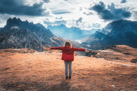 Young woman with raised up arms and mountains at sunset in autumn in Dolomites, Italy. Landscape with happy girl in red jacket, dramatic sky with clouds, orange grass and trees, high rocks in fallの写真素材