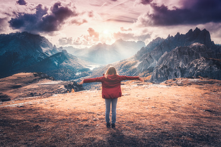 Young woman with raised up arms and mountains at sunset in autumn in Dolomites, Italy. Landscape with happy girl in red jacket, dramatic sky with clouds, orange grass and trees, high rocks in fallの写真素材