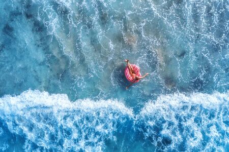 Aerial view of a young woman swimming with the pink donut swim ring in clear blue sea with beautiful waves at sunny day in summer. Tropical aerial landscape with girl, azure water. Top view. Travelの写真素材