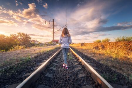 Standing young woman on the railroad at sunset in summer. Slim girl on the railway station. Rural industrial landscape with railroad, sky with colorful clouds. Railway platform. Transportation. Travelの写真素材