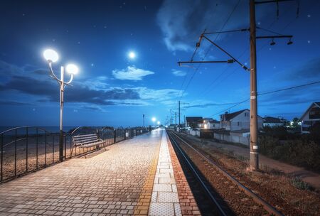 Beautiful railway station at night in summer. Starry sky over railroad at dusk. Industrial landscape with railroad, railway platform, city lights, buildings, blue sky with moon and clouds at twilightの写真素材