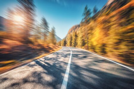 Mountain road in autumn forest at sunset with motion blur effect. Asphalt road and blurred background with orange trees, blue sky with sun in fall. Fast driving. Beautiful highway. Transportationの写真素材