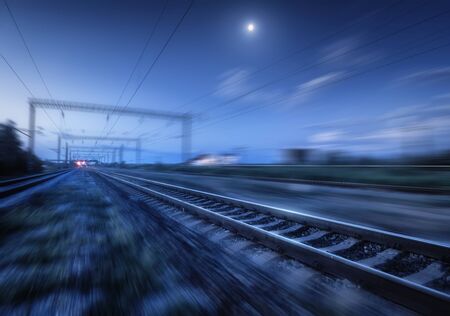Railroad and blue sky with moon and clouds at night with motion blur effect. Industrial landscape with railway station and blurred background at twilight. Railway platform in move. Transportationの写真素材