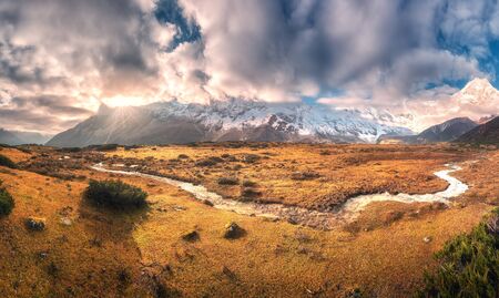 Panoramic landscape with mountains with snow covered peaks in clouds, sun, small river, orange...