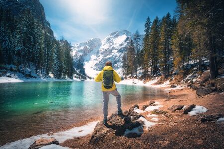 Sporty man with backpack is standing on the stone near lake with azure water at sunny day in autumn. Travel. Landscape with guy, reflection in water, snowy mountains, orange trees, blue sky in fallの写真素材