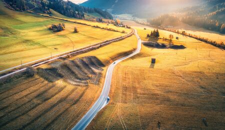 Aerial view of the road in mountain valley at sunset in autumn. Top view of asphalt roadway, railroad, hills with orange meadows, sunlight, yellow trees, buildings. Highway and fields in fall. Sceneryの写真素材