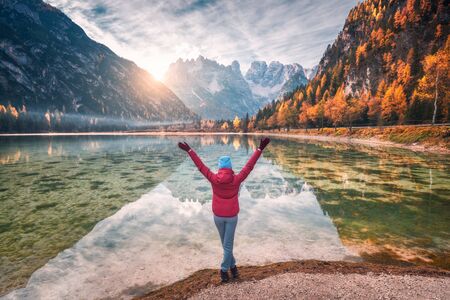 Happy young woman with raised up arms is standing near lake with clear water and mountains at sunset...