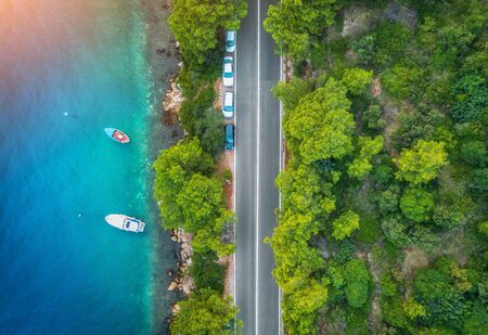 Aerial view of road in beautiful green forest and boats and yacht in the sea at sunset in summer. Colorful landscape with roadway, blue water, trees. Top view from drone of highway in Croatia. Travelの写真素材