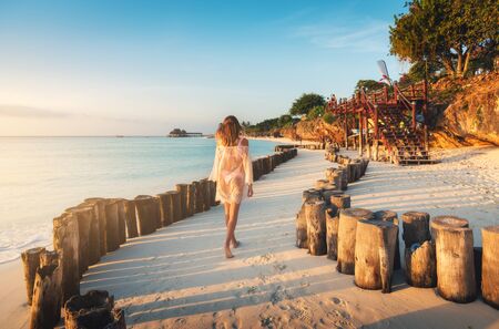 Beautiful young woman is walking on the sandy beach near blue sea at sunset. Summer travel. Tropical landscape with slim girl in white lace dress on the sea coast, wooden stumps, blue sky. Vacationの写真素材