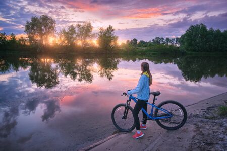 Woman with bicycle on the coast of the river at sunset in summer. Landscape with young girl with mountain bike, sandy beach, purple sky with colorful clouds reflected in water. Sport and travel. Cycleの写真素材