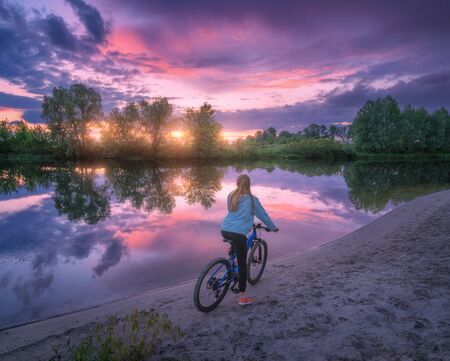 Woman with bicycle on the coast of the river at sunset in summer. Landscape with young girl with mountain bike, sandy beach, purple sky with colorful clouds reflected in water. Sport and travel. Cycleの写真素材