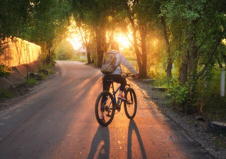Woman riding a bicycle on the rural road at sunset in summer. Colorful landscape with sporty girl with backpack riding a mountain bike, road, green trees and gold sunlight. Sport and travel. Cycleの写真素材