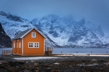 Orange rorbu on sea coast and snow covered mountain in fogの写真素材